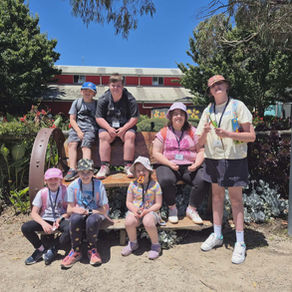 A group of eight children sit and stand on a wooden bench outdoors on a sunny day, smiling at the camera, with trees and a building behind them.