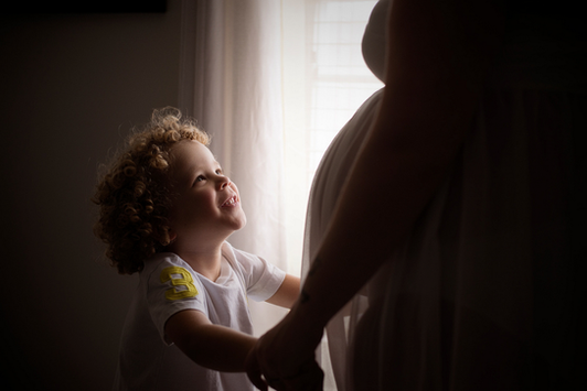 An excited brother-to-be holding his pregnant mother's hands standing next to light and airy window light.