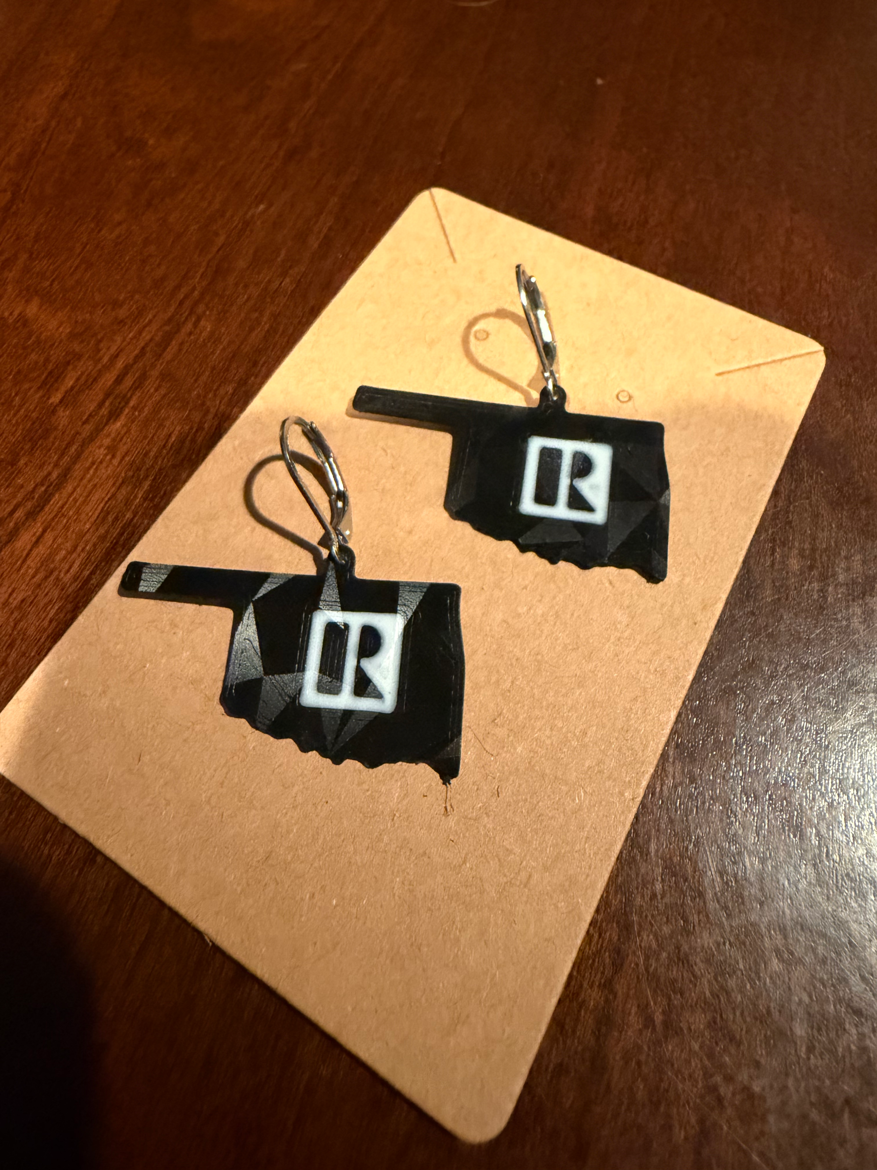 Black Oklahoma-shaped Real Estate Professional earrings with silver lever-back hooks on a display card