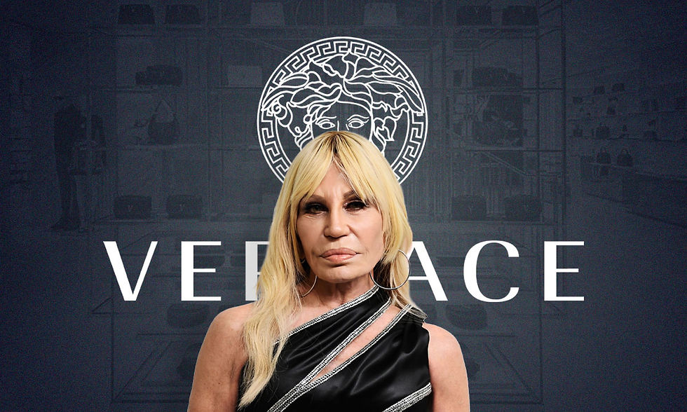 Donatella Versace con vestido negro ante el logo de Versace, con expresión seria, en fondo oscuro.