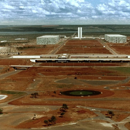 Foto aérea histórica do Eixo Monumental em Brasília no início dos anos 1960. O cenário é marcado pela terra vermelha batida do cerrado, evidenciando o impacto geográfico e social da construção civil-militar da nova capital. No centro, estende-se a Esplanada dos Ministérios deserta, com os blocos ministeriais em fase de conclusão e as duas torres verticais brancas do Congresso Nacional ao fundo. À esquerda, avista-se a estrutura esquelética de concreto da Catedral Metropolitana. O céu ao fundo está carregado de nuvens claras sobre o horizonte plano do Planalto Central.