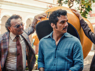 Cena do filme O Agente Secreto com o ator Wagner Moura, que interpreta o protagonista, no centro da imagem, ao lado de outros atores do filme em um lugar aberto na frente de um telefônico público (orelhão).