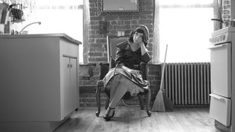 Thoughtful seated portrait with vintage styling, suggesting inner reflection in a creative space Emily Carmen is seated in front of a brick wall leaning her head on her hand