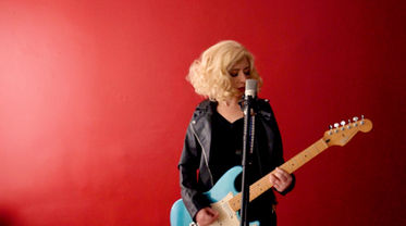 Tight frame of hands strumming a turquoise Fender Stratocaster, part of a color-rich sequence symbolizing artistic expression in contrast to black-and-white domestic scenes in the video.