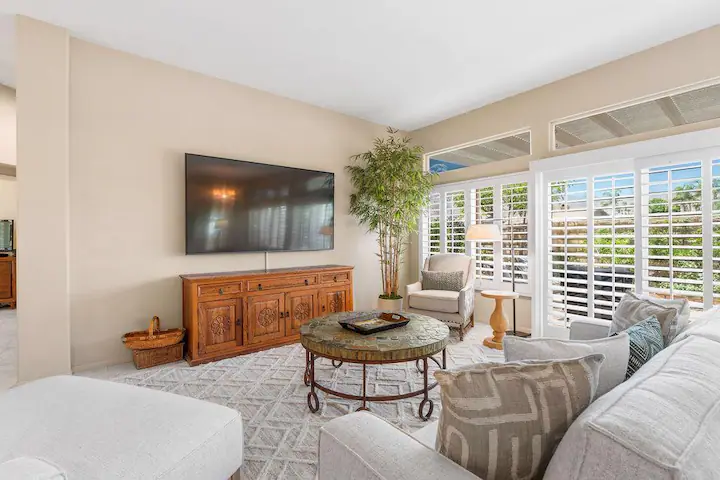 Living room with comfortable couches, flat screen tv, and large shuttered windows to backyard with private pool