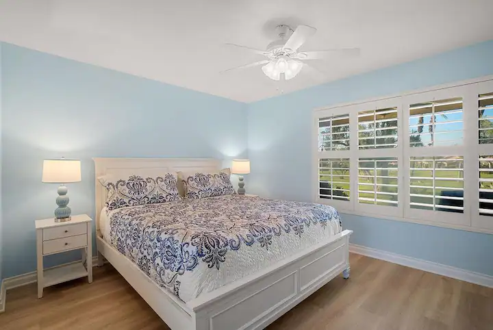 Master bedroom in Palm Desert vacation rental home with blue walls and king bed, two night stands with lamps, and large shuttered windows looking over a golf course