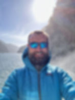 Smiling man in a blue jacket standing on a boat at Milford Sound, New Zealand, with sunlight and waterfall spray in the background.