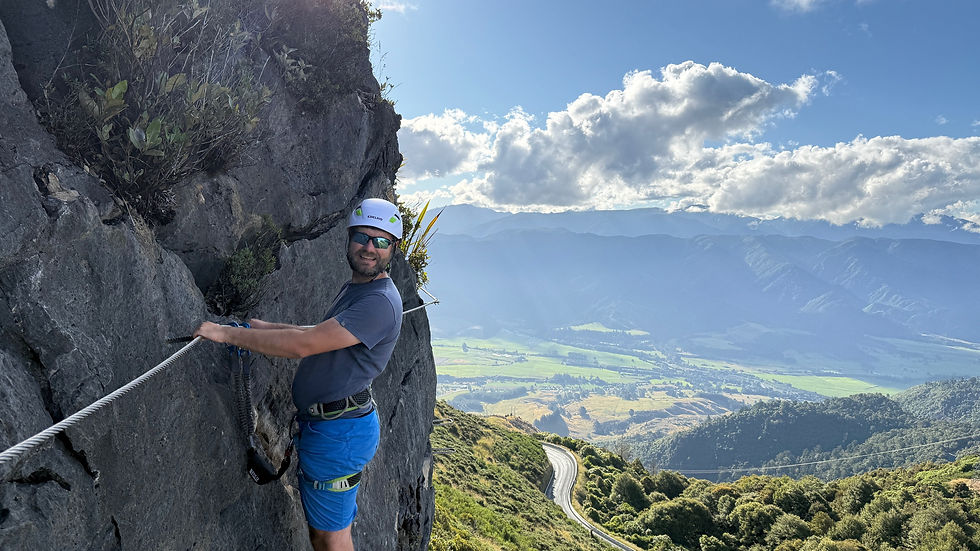 Man climbing a rock face with safety gear on the Cable Bay Via Ferrata track, overlooking valleys and mountains in Nelson, New Zealand.