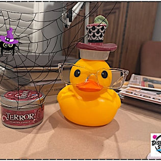 Yellow rubber duck wearing glasses and a tiny porcelain hat sits beside a candle labeled “Terror,” on a brown paper-covered desk with a spiderweb and the Disorderly Studio logo overlay.
