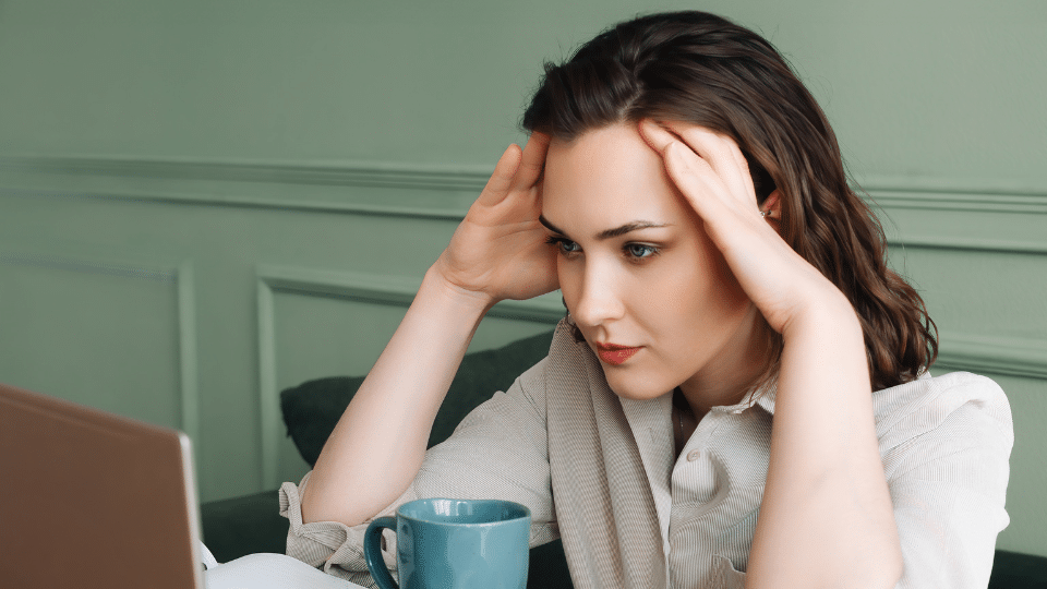 A woman looks stressed, holding her head while sitting at a desk with a laptop and a blue mug. The background is light green walls.