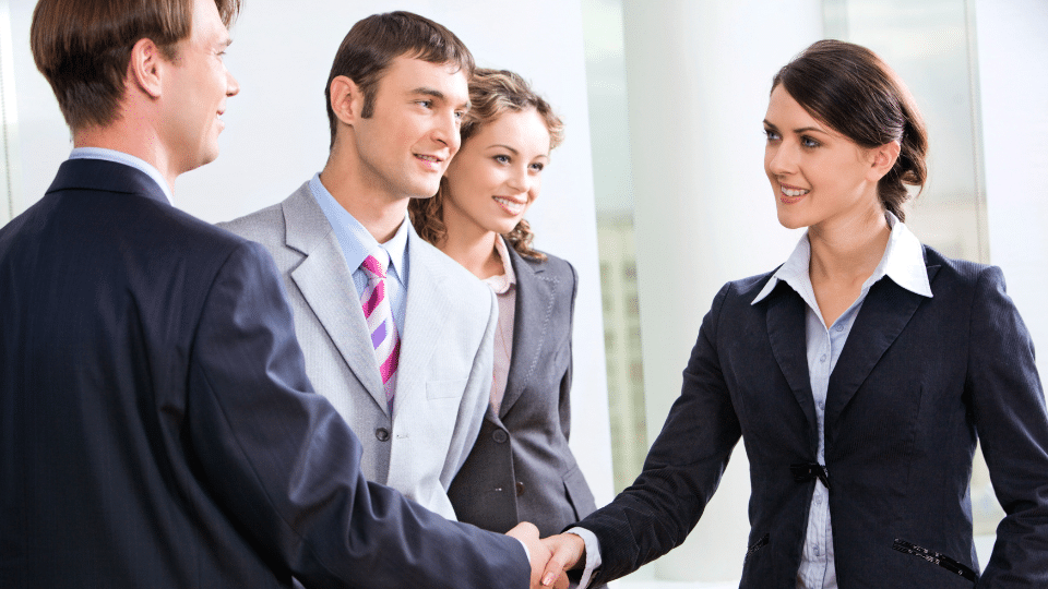 Four people in business attire smile in an office setting. Two in front are shaking hands, suggesting a positive, professional interaction.