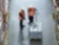Two warehouse workers in safety vests and hard hats reviewing paperwork beside a pallet of shrink-wrapped canned goods in a storage aisle.