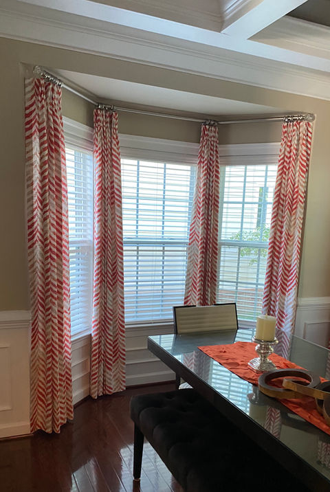 Dining room bay window with white horizontal blinds and red and white patterned custom curtains on a curved rod, framing a dark dining table with an orange runner, illustrating bay window treatments by Mill End Shops Rockville MD