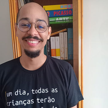 Foto de Ivan, um homem negro, careca, de barba e bigode, utilizando óculos. Ivan usa uma camisa preta com os dizeres "um dia, todas as crianças terão uma educação de qualidade" e "ensina BRASIL". Ao fundo, há uma estante com livros.