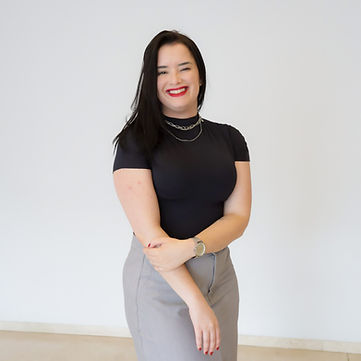 Foto de Fabiane Macedo, uma mulher branca de cabelos lisos. Fabi usa uma camisa preta de mangas curtas, uma saia cinza, relógio e colares, além de batom e esmalte vermelhos. Ela encontra-se com os braços em frente ao corpo, com a mão esquerda posicionada sobre o cotovelo direito. Ao fundo, há uma parede cinza claro.