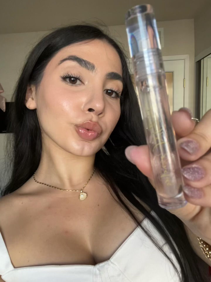 Woman with long dark hair holds a clear lip gloss tube, posing confidently in a white top. Neutral-tone background, kissy lips.