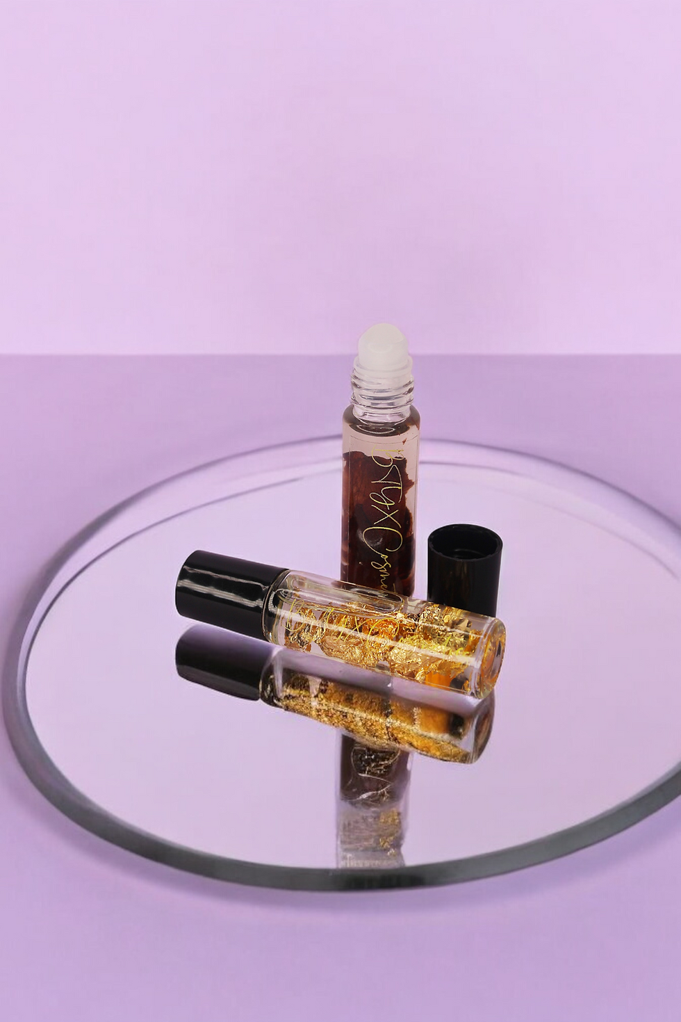 Two lip oil bottles on a round mirror with a purple background. One bottle is upright, the other lying down, showing gold flakes inside.