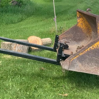 Clamp-on bucket forks on a tractor in the grass near chopped wood.