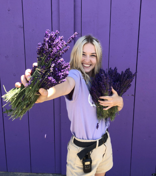 A smiling woman in a lavender shirt holds bunches of lavender flowers against a purple wall. She's wearing beige shorts and a black belt.