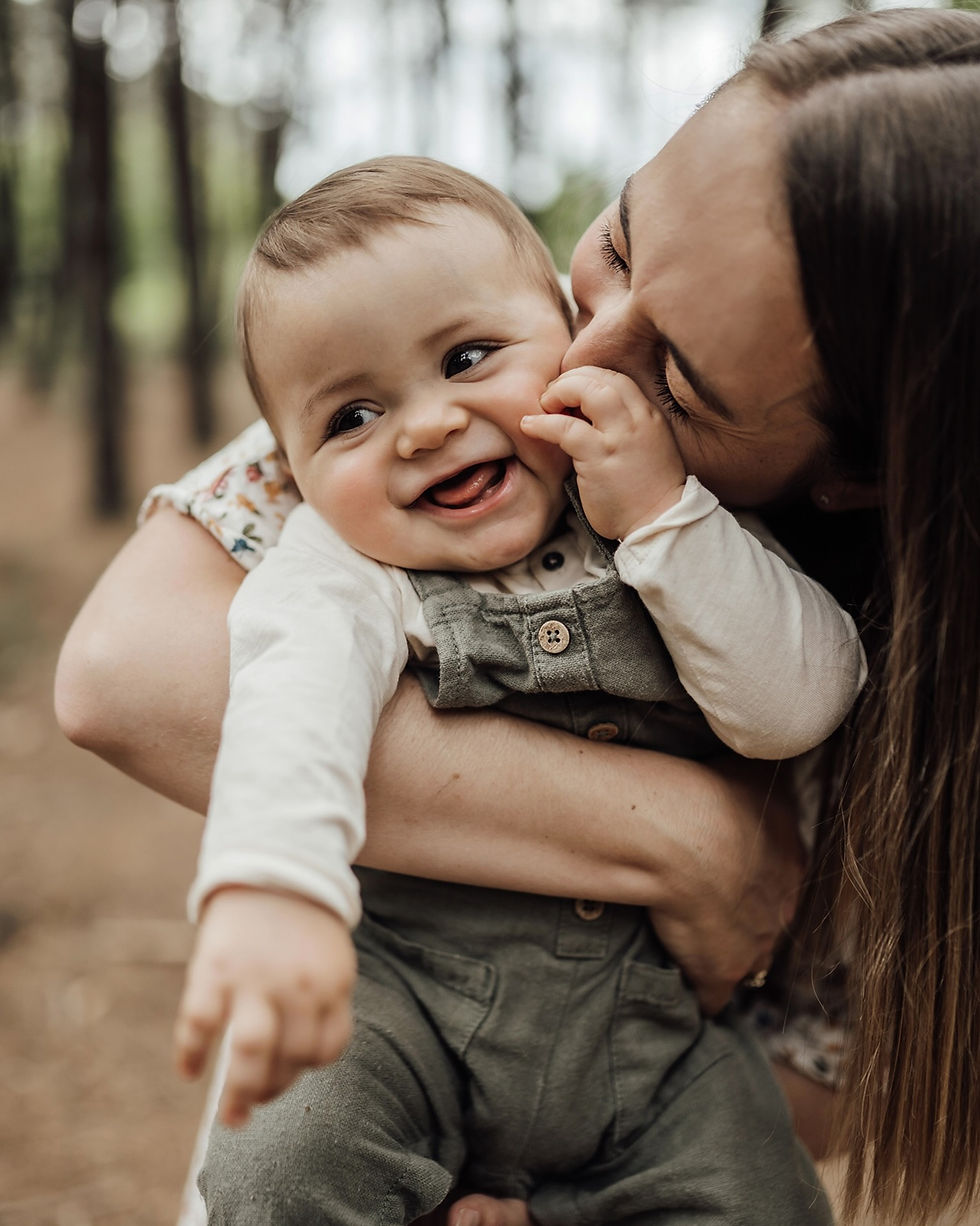 Mère embrassant son bébé souriant dans une forêt, portrait tendre et joyeux.