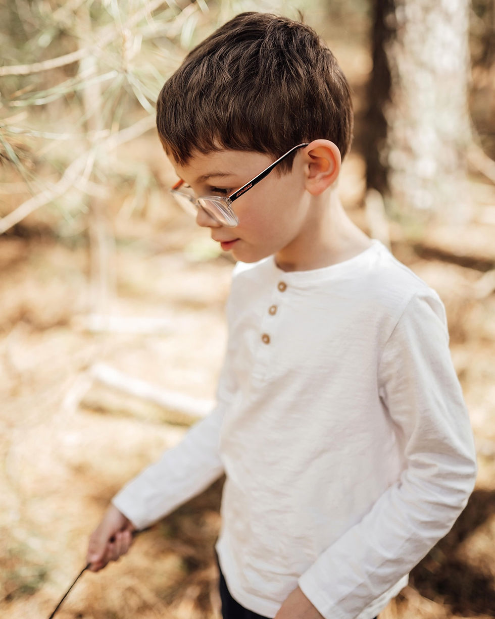Garçon en chemise blanche et lunettes, regard baissé, tenant un bâton dans une forêt aux tons dorés.