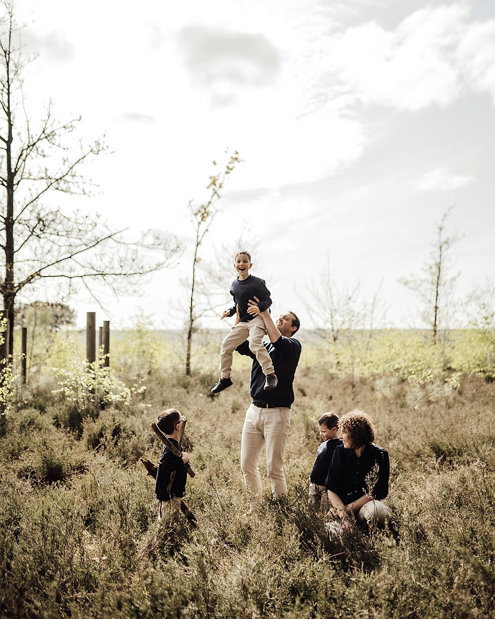 Famille souriante dans une prairie, un père soulève son fils, mère et deux enfants près d’eux sous un ciel clair.
