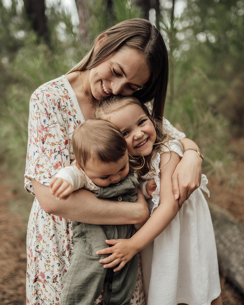 Mère souriante enlacée avec deux jeunes enfants en forêt, l’une en robe blanche, l’autre en salopette verte.