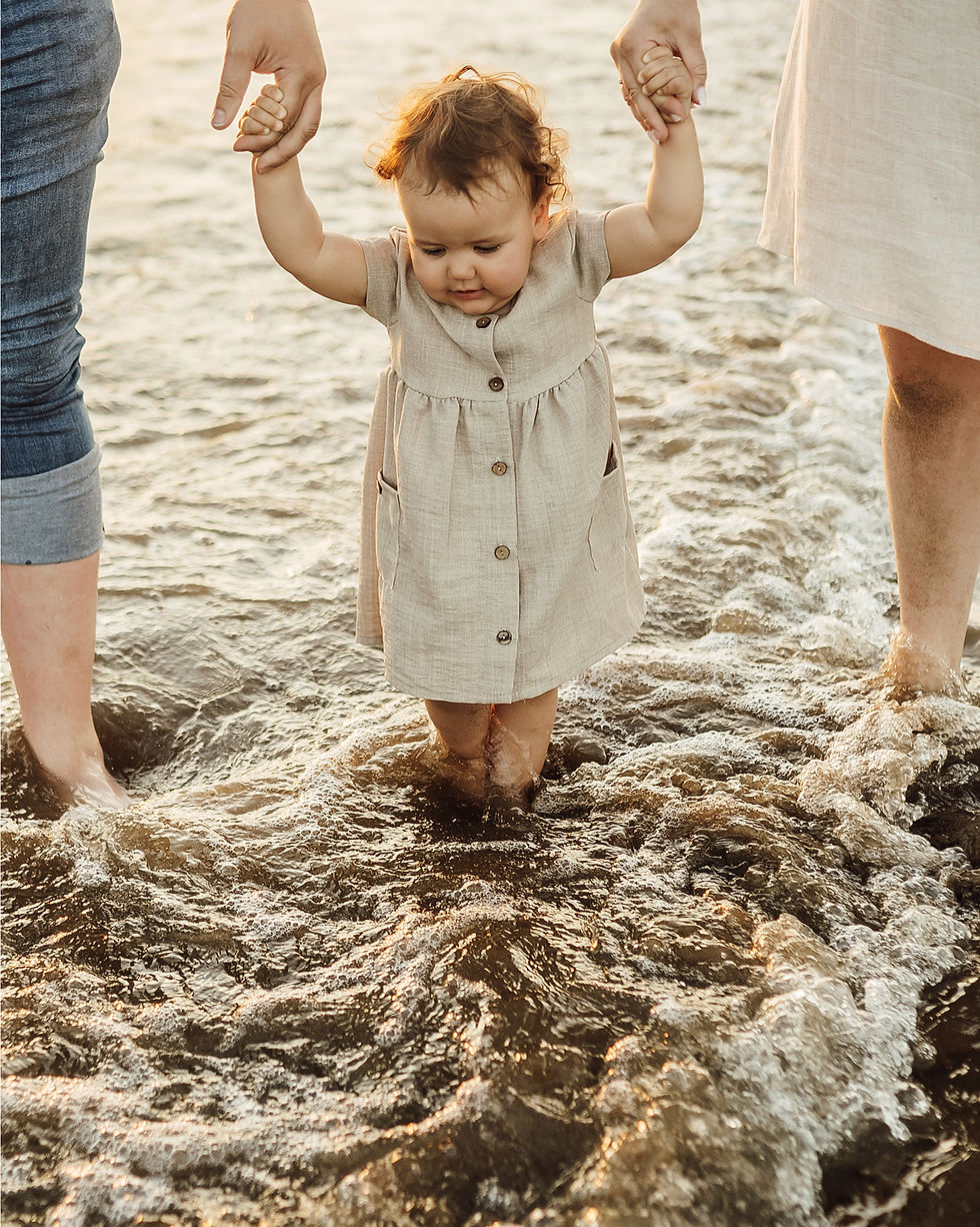 Bébé en robe beige, tenu par deux adultes, avance dans l’eau au bord de mer, air concentré, lumière dorée.