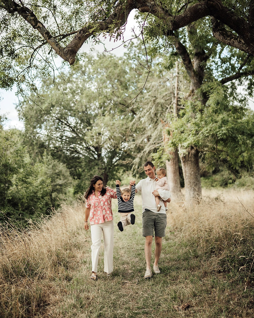 Famille de quatre sur un sentier boisé, parents souriants portant un bébé et un enfant qui se balance joyeusement.