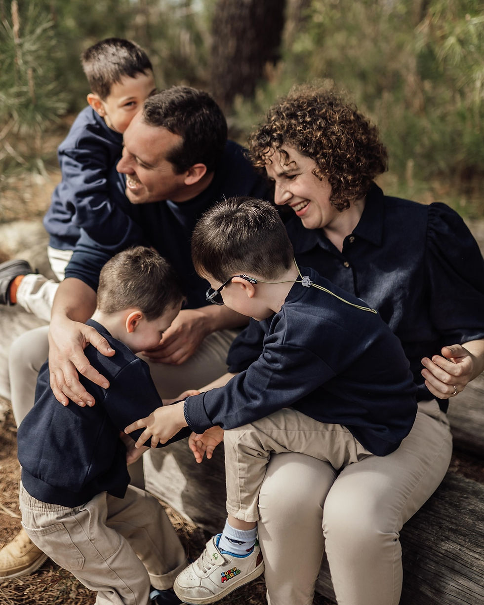 Famille souriante dans la forêt, parents assis sur un tronc, deux enfants en bleu se serrent; chaussure Super Mario visible.
