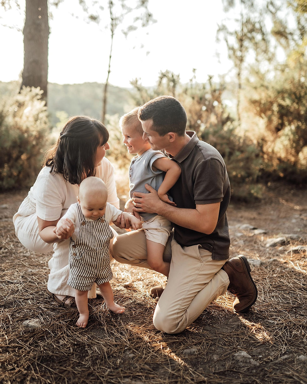 Famille de quatre agenouillée en forêt, parents souriant aux deux jeunes enfants, lumière dorée, ambiance tendre.