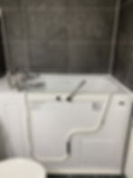 A bathtub shell in a bathroom with grey tiles and a toilet seat