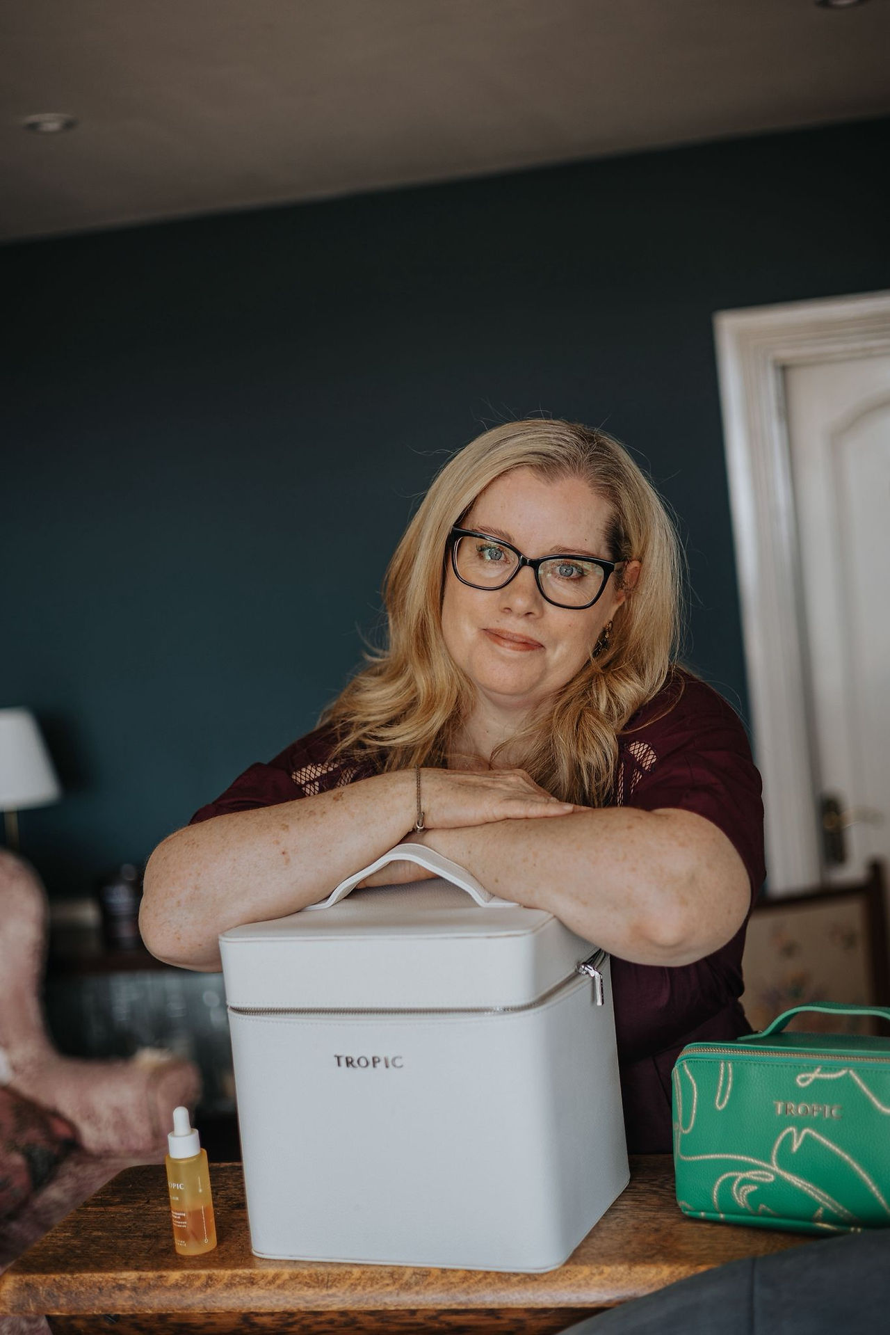 Blonde woman with glasses, smiling and leaning on a white box.