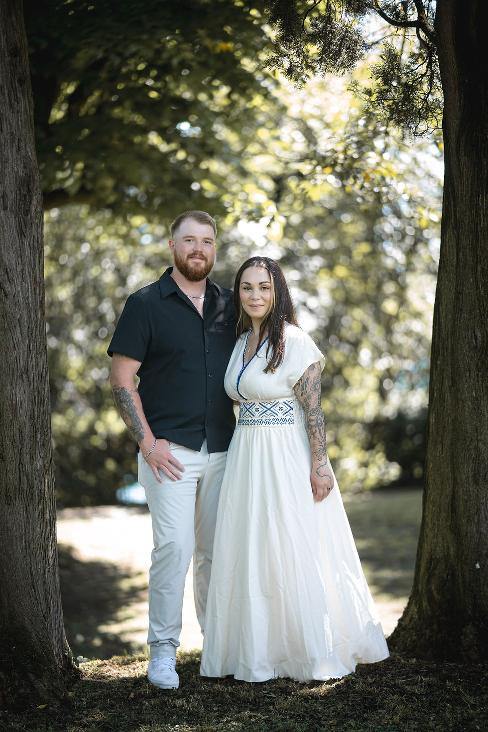 A couple stands between trees, smiling. The man wears a black shirt and white pants; the woman, a white dress. Sunlight filters through leaves.