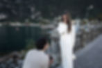Man kneels, holding ring, proposing to woman in white. She appears surprised, covering mouth. Lakeside setting with mountain backdrop.