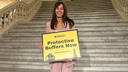 Lois Bower-Bjornson holding a sign stating "Protective Buffers Now!" against an ornate staircase.