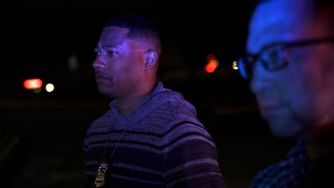 Two plainclothes police officers, bathed in the red and blue of their siren lights, watch attentively.