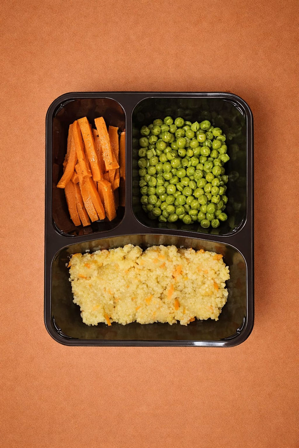 Meal with carrots, peas, and millet in a black tray on a brown surface