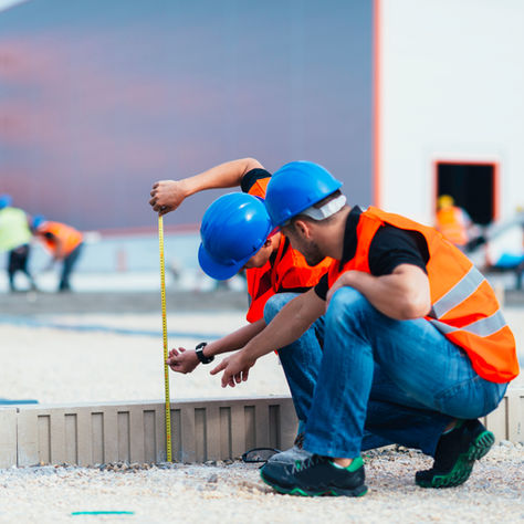 Two construction workers in blue hard hats and orange vests kneel measuring a curb with a tape measure at a building site.