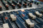 Close-up of a sound mixing board with various sliders and knobs on a dark background. One orange light is lit, indicating an active channel.