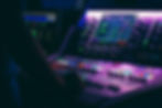 A person adjusts controls on a colorful, illuminated audio mixing console in a dimly lit studio, with digital screens displaying data.