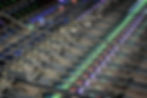 Audio mixing console with illuminated faders and buttons in shades of blue, green, and red. Displays show active channels in a studio.