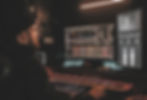 Man in a beanie working at a music studio. He's focused on mixing tracks on dual monitors with colorful audio waveforms. Dim lighting.