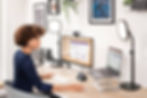 Woman working at desk with dual monitors, laptop, and microphone. Bright lights, modern decor, and framed art in the background.