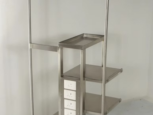 Stainless steel hospital trolley with drawers and wheels placed beside a hospital bed in a clinical ward setup.