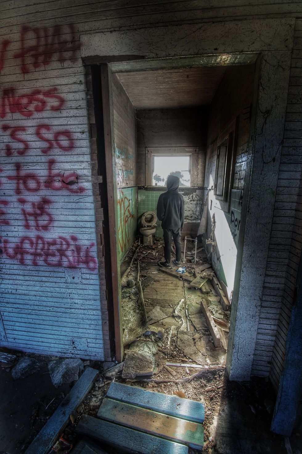 A small figure wearing all black standing with their back to the camera looking out of a brightly lit open window in a decrepit bathroom.