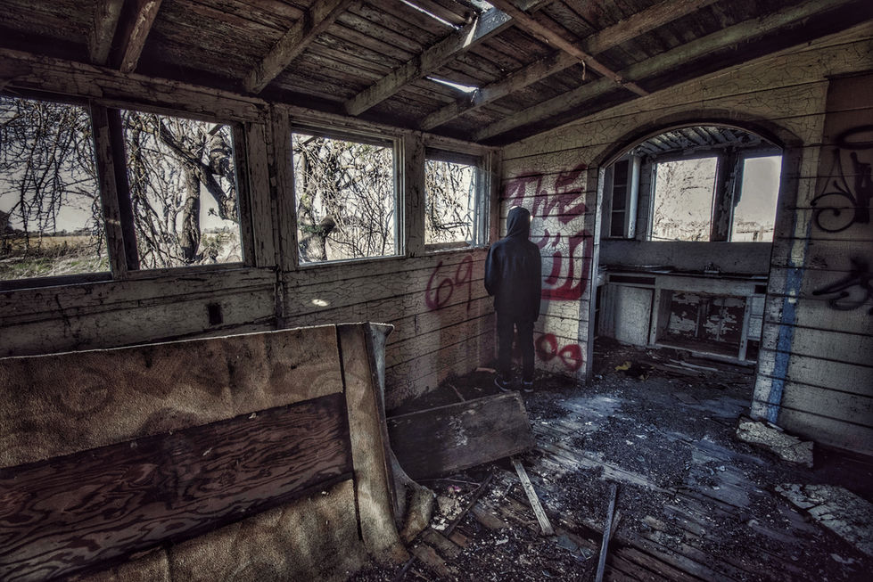A small figure wearing a black hoodie and black pants standing with their back to the camera in the corner of an abandoned and broken down house. "The Kid" spray-painted on the wall.