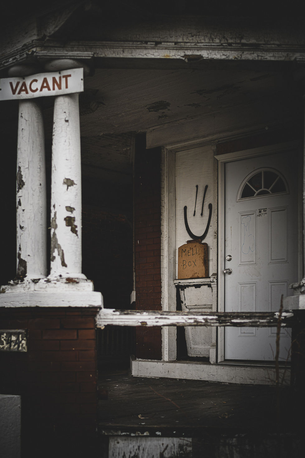 Porch of an abandoned house with a vacant sign and a spray-painted smiley face next to the front door.