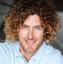 Raphael - headshot of a male actor with long curly hair, groomed beard, and wearing a blue shirt, represented by ROC Talent Agency UK