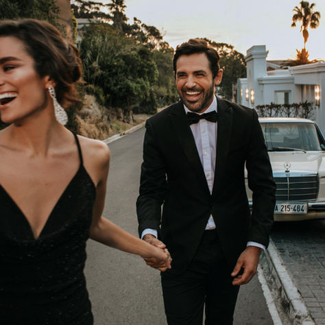 A couple smiling running towards the camera hand in hand, dressed in evening wear, and showcasing authentic connections.